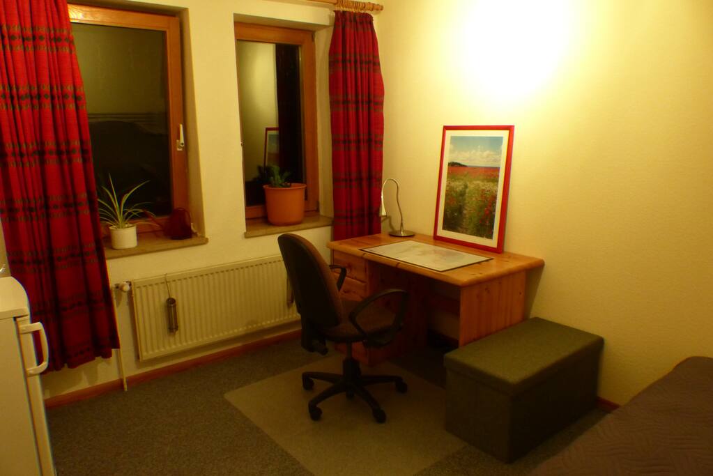 Blick von der Tür auf die Fensterfront, den Schreibtisch mit Stuhl und die Sitztruhe