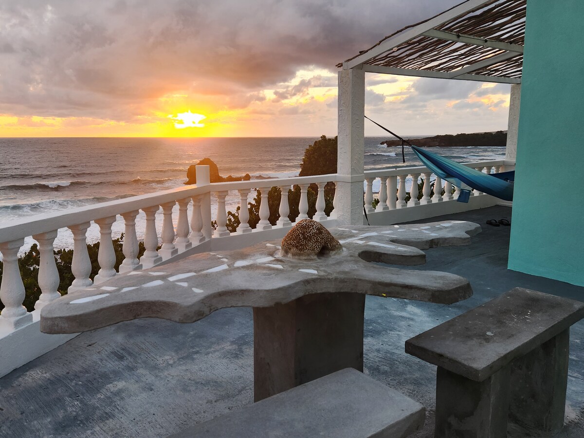 A concrete table and benches are positioned on a private terrace, overlooking the ocean. The scene is illuminated by a vibrant sunset, with orange and yellow hues reflecting on the water. A hammock is visible, gently swaying in the breeze.
