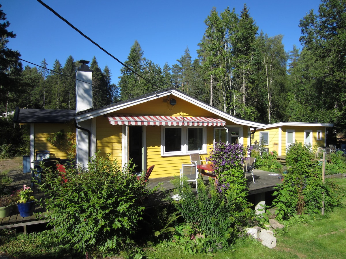 A vibrant yellow cabin is surrounded by greenery, featuring a spacious deck with multiple chairs and a table for outdoor seating. A striped awning provides shade over the front entrance, while a separate structure can be seen in the background.