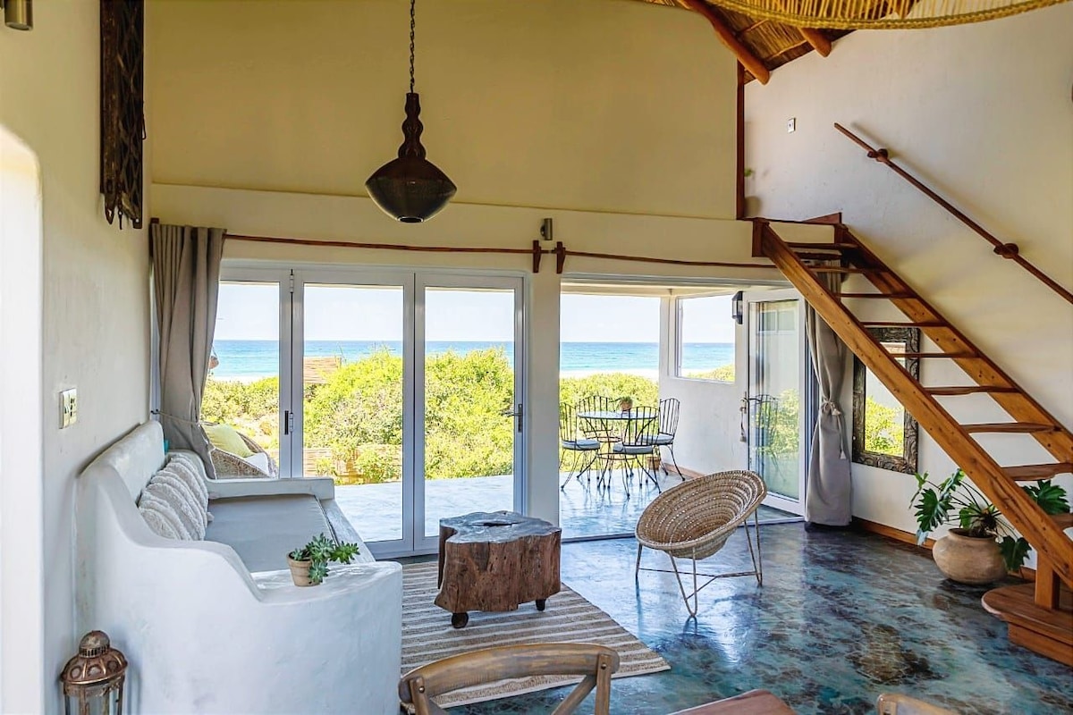 The spacious living area features a white sofa and a wooden coffee table, complemented by large glass doors that open to a sea view. A staircase leads to an upper level, while outdoor seating can be seen through the doors.