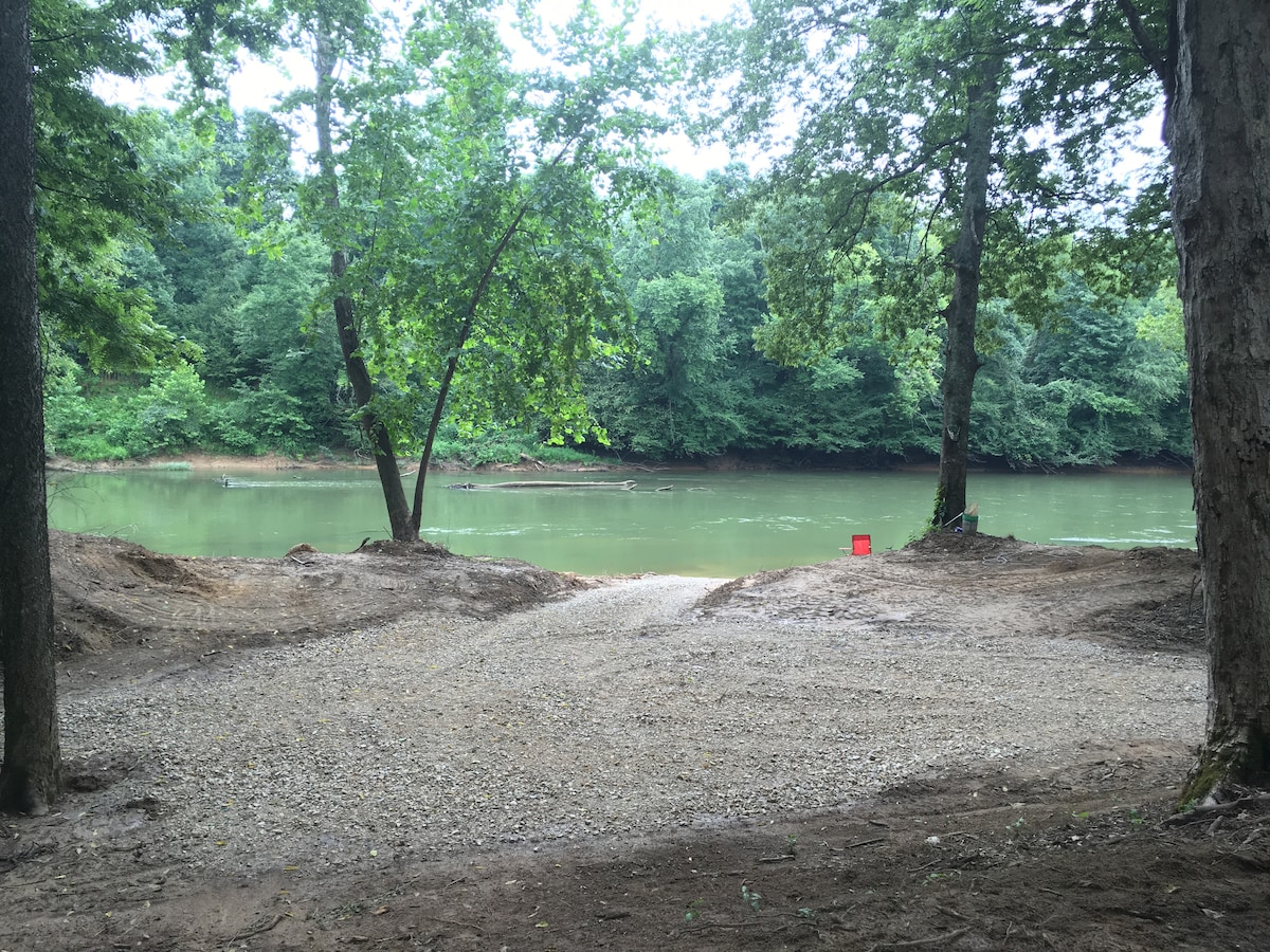 A gravel path leads to the Black River, framed by lush greenery and trees. The calm water reflects the surrounding vegetation, with a solitary red bucket positioned nearby, adding a pop of color to the serene environment.