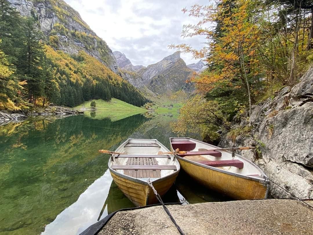 Two wooden boats are moored at the edge of a serene lake, reflecting the surrounding mountains and autumn-colored trees. The calm water mirrors the vibrant foliage, creating a peaceful scene. The area is framed by lush greenery and distant rolling hills.
