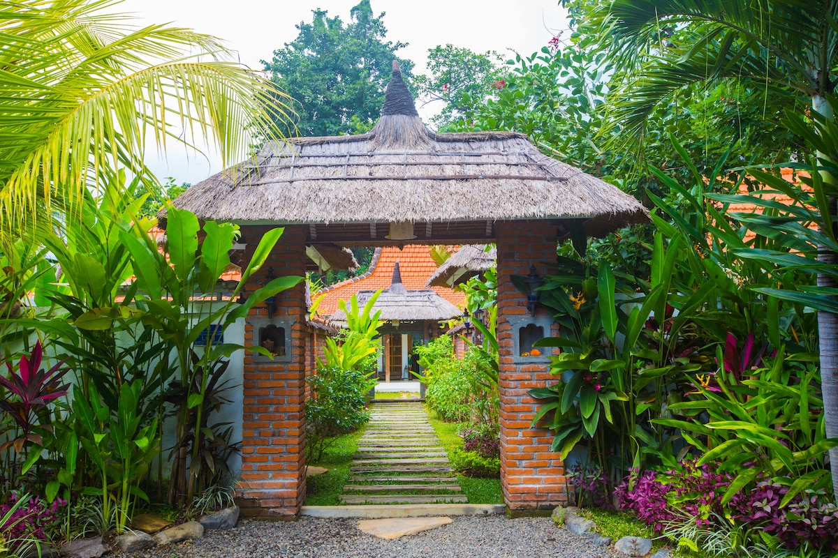A welcoming entrance is framed by lush tropical plants and vibrant flowers. A path leads through a rustic archway to the main building, which is topped with a traditional thatched roof. The tranquil atmosphere invites exploration of the surrounding garden.