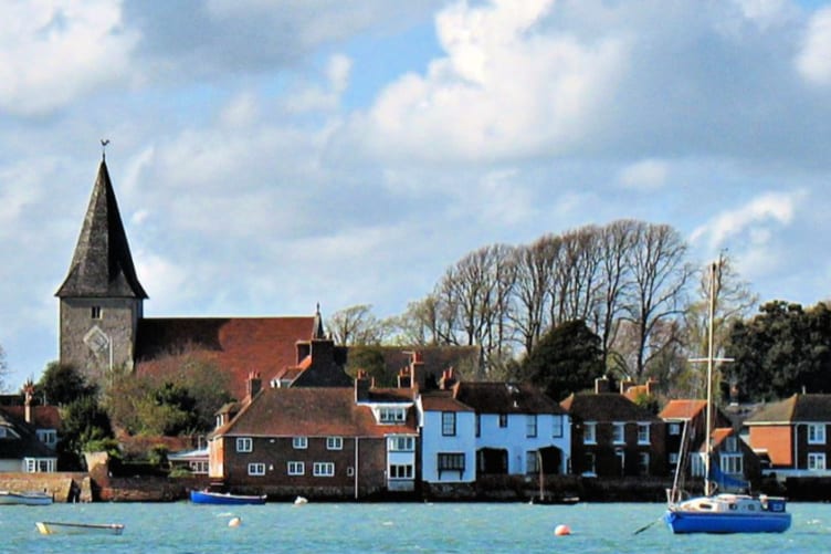A charming coastal scene depicts a cluster of traditional homes with red roofs lining the shore. A tall spire rises from a church, and anchored boats bob gently in the water. The backdrop is adorned with trees, under a sky filled with fluffy clouds.