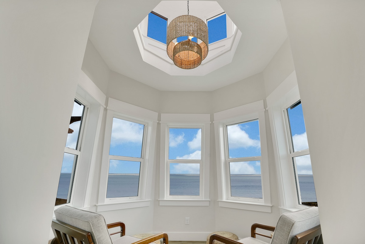 A bright seating area features large windows offering views of the beach and sky. A pendant light hangs from the octagonal ceiling, illuminating the space. Cozy lounge chairs are placed for relaxation, enhancing the serene atmosphere of this waterfront retreat.