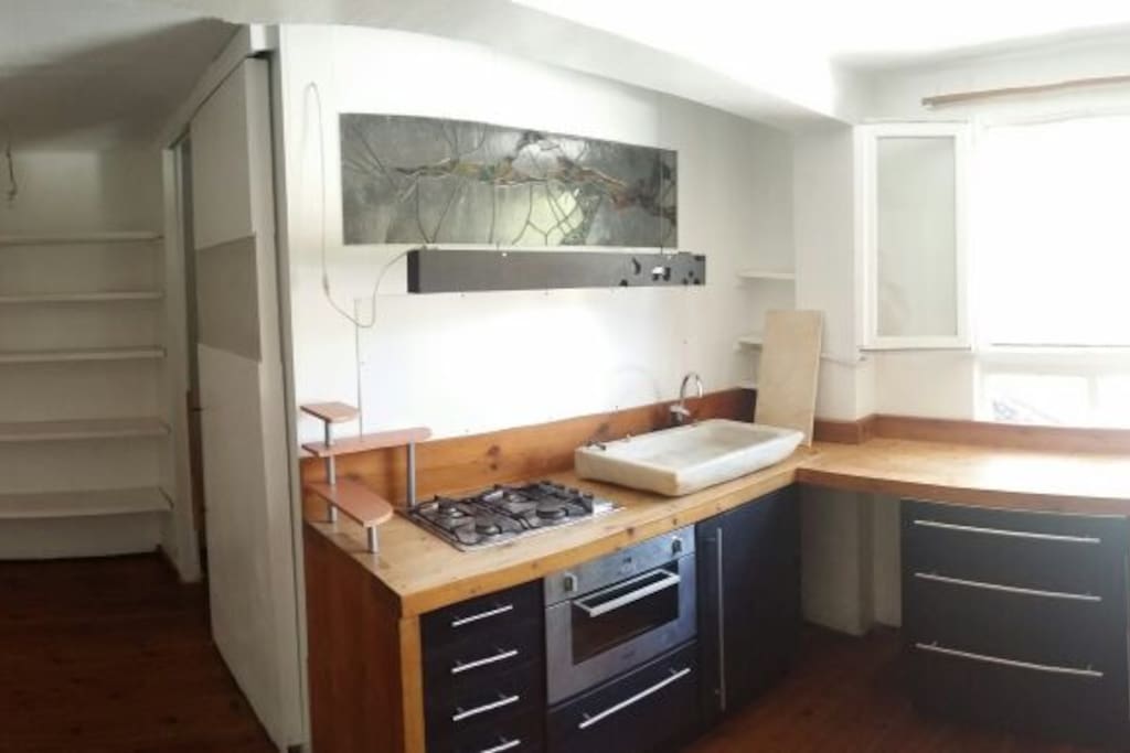 de izquierda a derecha: puerta de entrada, armario con estanterias, puerta corredera del baño, cocina de madera, fogones de gas, horno electrico, armarios de cocina negros, pica de cocina de marmol blanco, ventana con vista a terreno verde