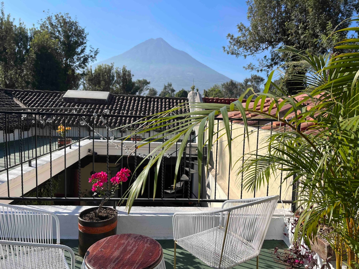 A rooftop terrace features two white woven chairs and a circular wooden table. Lush green plants are present, with bright pink flowers in a pot. In the background, a clear view of a volcano can be seen against a blue sky, adding to the serene atmosphere.