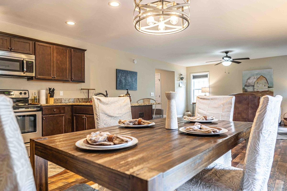 A spacious dining area features a rustic wooden table set with elegant plates and napkins. Stylish chairs surround the table, while a modern chandelier provides overhead lighting. The kitchen, visible in the background, showcases dark cabinetry and stainless steel appliances.