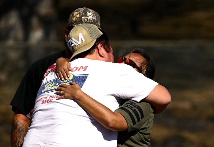 Tres personas paradas bajo el sol se abrazan y sus rostros no se alcanzan a ver por el abrazo. Uno lleva una gorra hacia atrás.