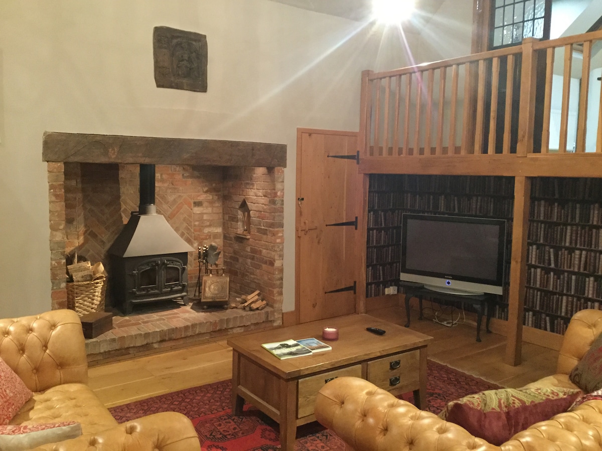 A spacious lounge is highlighted by a charming inglenook fireplace made of brick, accompanied by a cozy seating area featuring leather sofas. A wooden coffee table sits centrally, while a television is positioned against a wall adorned with shelves of books, adding character to the space.