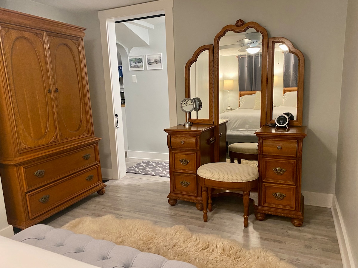 A charming dressing area features an ornate wooden vanity with a mirror and a coordinating stool. A spacious armoire stands nearby, showcasing vintage design elements. Soft natural light softly illuminates the room, enhancing the light color of the walls and flooring.