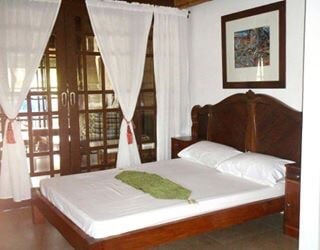 A sleeping area is presented with a wooden bed frame and a light-colored mattress. Sheer curtains frame large glass doors, offering a view of the outdoors. A green accent piece is neatly placed on the bed, and a piece of art decorates the wall.