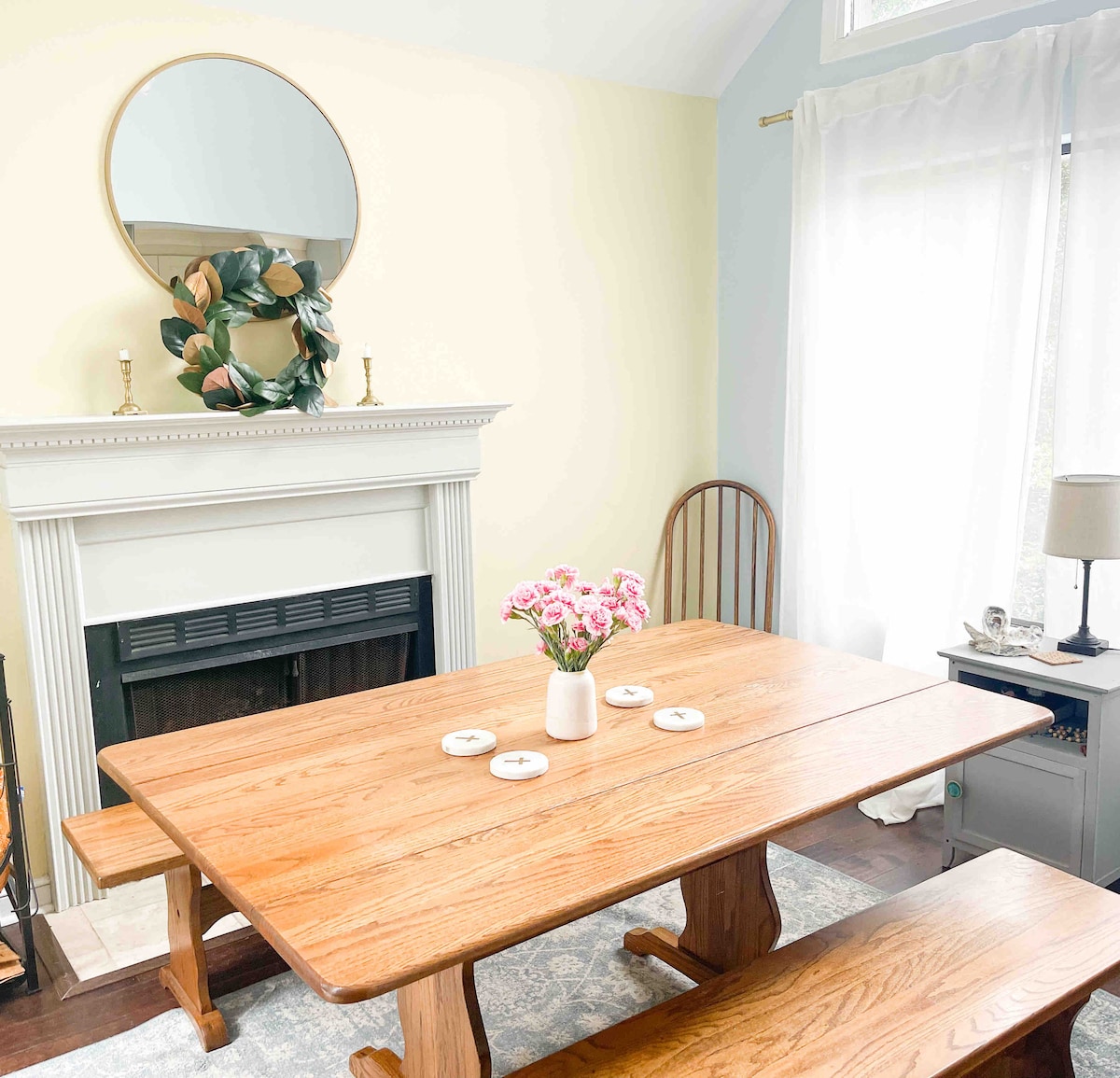 A wooden dining table is centered under a circular mirror, complemented by a floral centerpiece. The space features a cozy fireplace and a chair placed nearby. Light filters through sheer curtains, creating a bright atmosphere in the room.