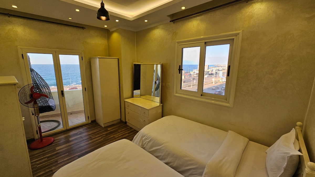 A well-lit bedroom features two spacious beds, each with crisp white linens. Natural light floods the room through large windows, offering a view of the sea. A dresser and mirror are positioned against an accent wall, while a ceiling fan enhances air circulation.