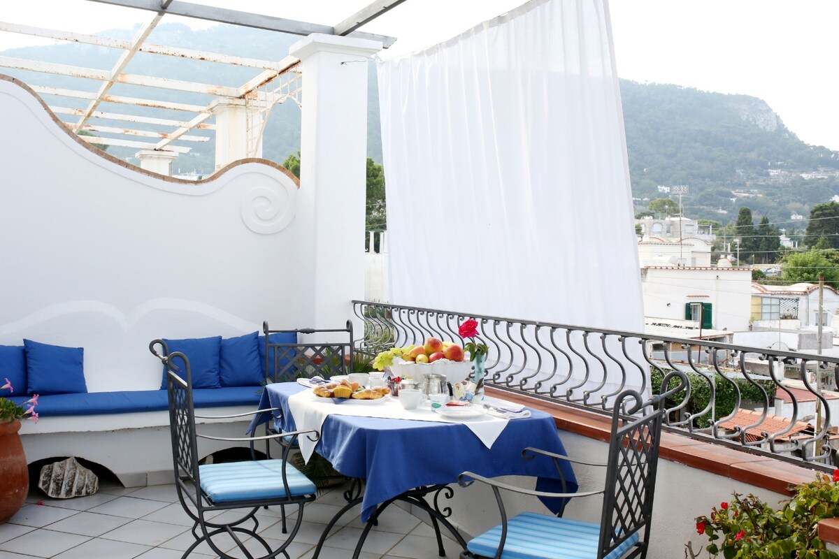 A terrace area features a round table set with a white tablecloth, accompanied by dishes of food and fresh fruit. Metal chairs with blue cushions surround the table, and a built-in seating area with blue cushions is visible. A white fabric drape adds privacy, with hills in the background.