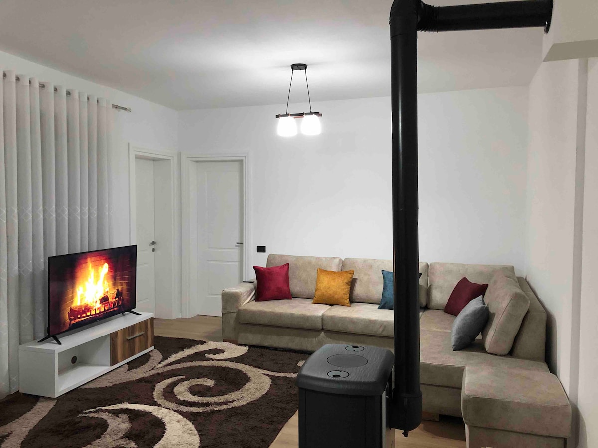 A spacious living area features a soft sectional sofa adorned with colorful cushions. A modern television is mounted on a white unit, displaying a cozy fireplace scene. A textured area rug covers the floor, complementing the light walls and providing a welcoming ambiance.