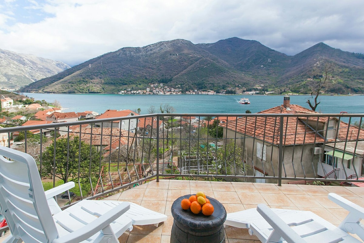 A balcony overlooks the scenic Boka Kotor Bay, framed by mountains in the background. Two white lounge chairs are positioned for relaxation, alongside a small table displaying a bowl of oranges. The tile floor and railing complete the inviting outdoor space.