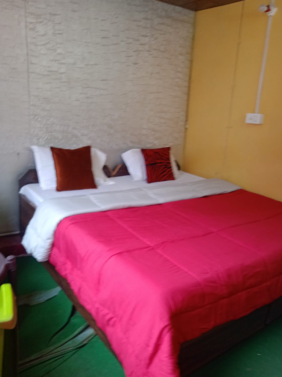 A spacious bed is beautifully made with a vibrant red blanket and accented by decorative pillows in shades of orange and rust. The room is adorned with textured white walls and a warm yellow accent wall, providing a cozy atmosphere.