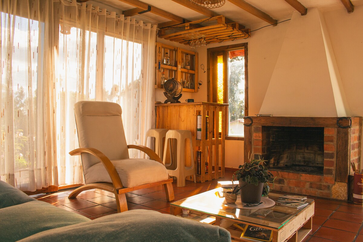 A cozy living area features large windows that allow natural light to fill the space. A comfortable armchair is positioned nearby a rustic fireplace, while a wooden bar unit and a glass coffee table are also present. The terracotta floor adds warmth to the setting.
