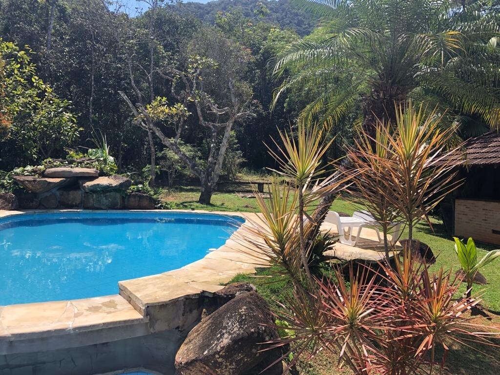A well-maintained outdoor swimming pool is surrounded by lush greenery and rocks. Vibrant plants add color, while poolside lounge chairs are positioned for relaxation. Distant trees and hills create a serene backdrop, enhancing the natural setting.