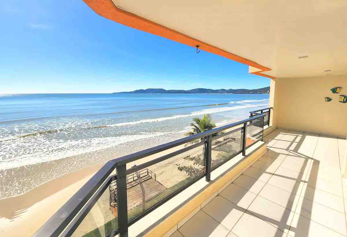 A spacious balcony overlooks a sandy beach and the ocean, with gentle waves lapping at the shore. The area is bathed in natural light, and the expansive glass railings provide an unobstructed view of the coastline and distant mountains.