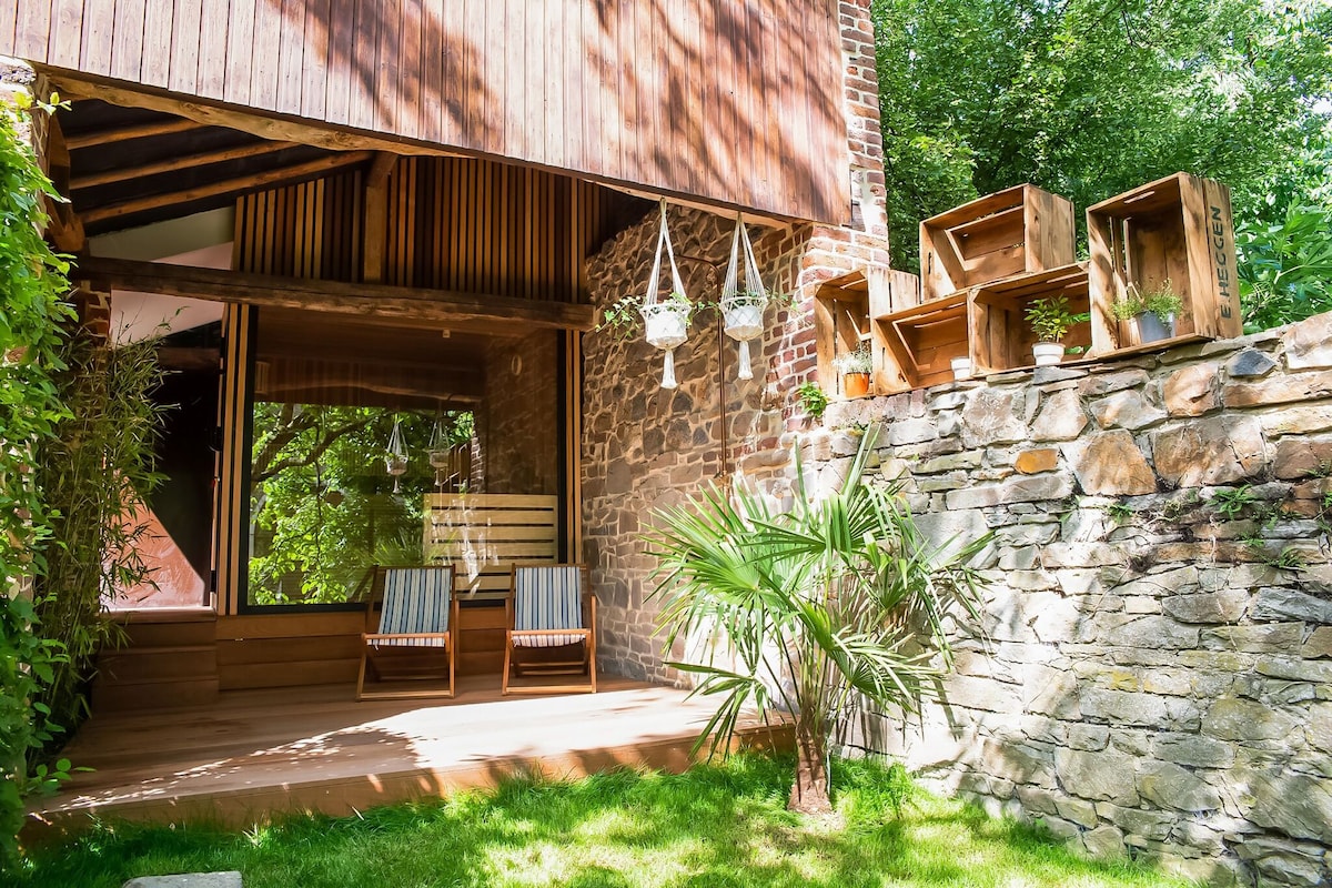 A private terrace is featured, bordered by stone walls and greenery. Two lounging chairs are positioned on a wooden deck, with potted plants hanging above and placed on shelves. Natural light filters through large glass doors, offering a seamless connection to the outdoor space.
