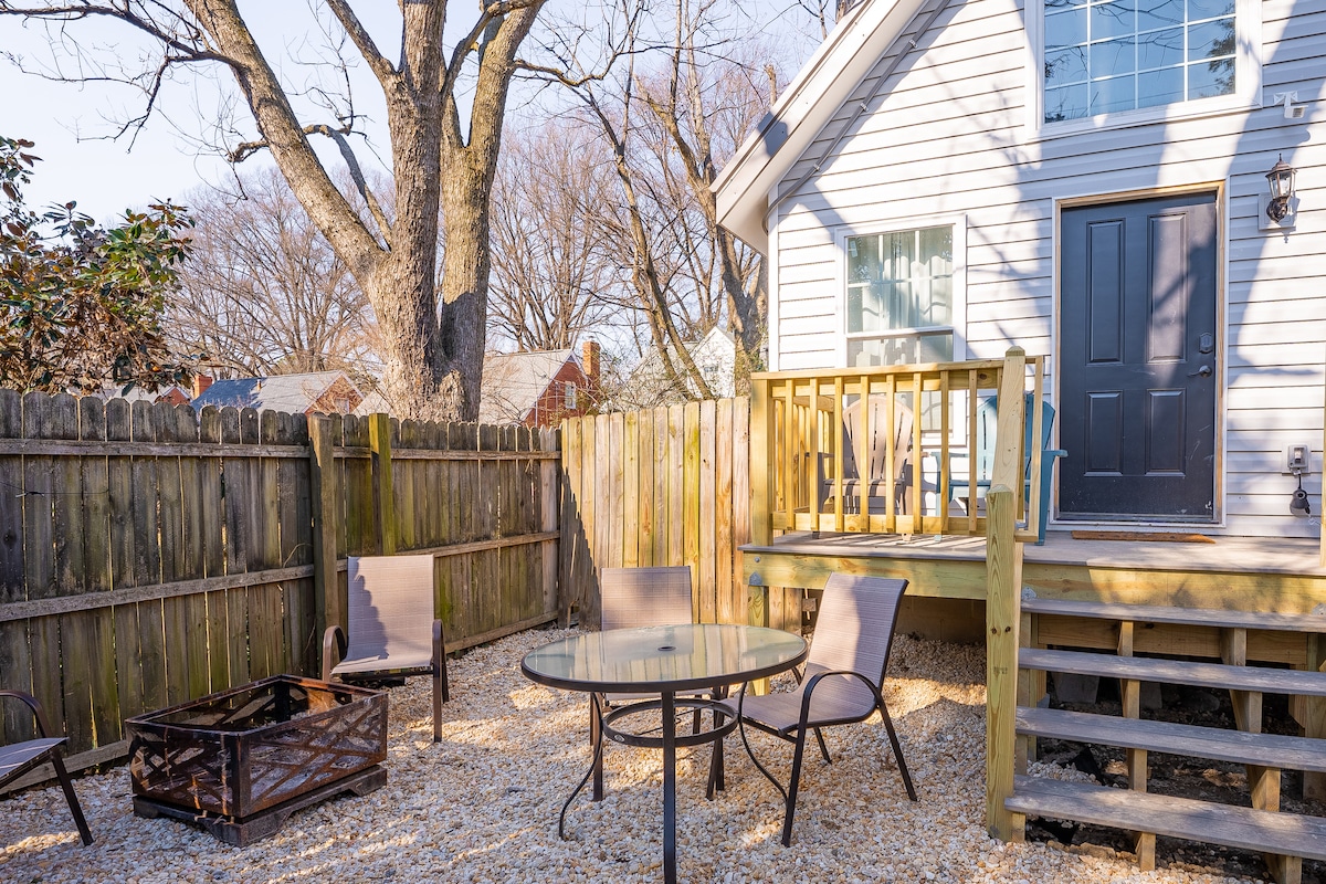 A private backyard space features a gravel patio with a circular table surrounded by four chairs. A fire pit is placed on the side, and wooden steps lead to the entrance of the guest house. The fenced area offers shade from a large tree.