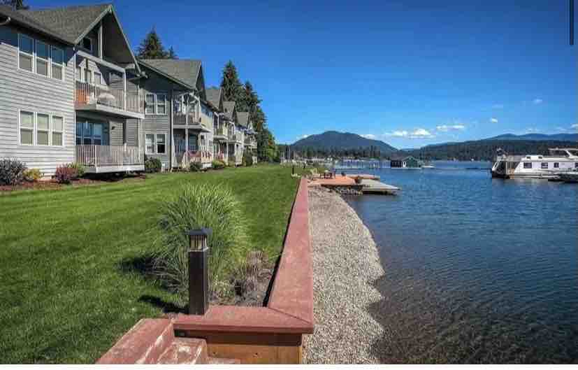 The image shows a well-maintained grassy area leading to a calm waterfront. Several condominium units are visible, each with balconies. A sandy shoreline edges the clear water, with boats docked nearby and mountains in the distance under a bright blue sky.