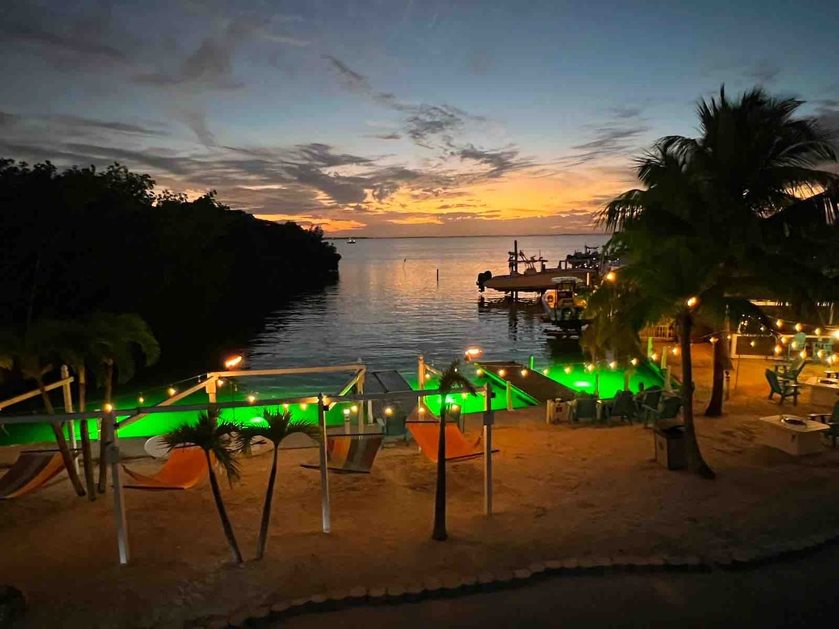 The view captures a serene bay at sunset, with vibrant colors reflecting on the water. A lit pool area is framed by palm trees, and hammocks are positioned nearby. A boat ramp is visible, with social seating arranged along the shore, enhancing the tranquil setting.