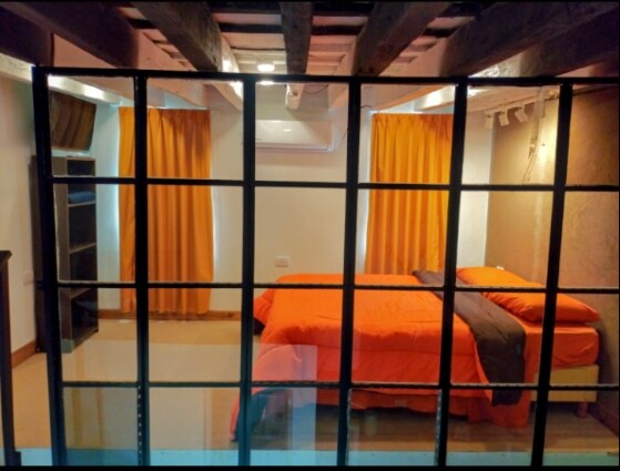 The room features a spacious bed with an orange bedspread and dark gray accents. Large windows with bright yellow curtains frame the space, and wood beams are visible on the ceiling, adding a touch of rustic charm. A shelf is positioned against one wall.
