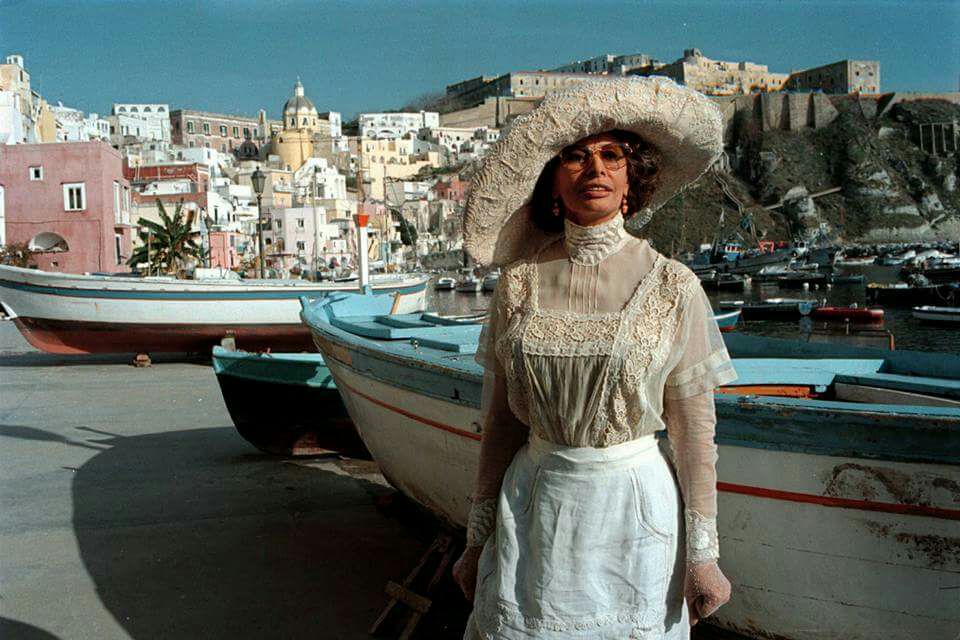 A figure in a large hat stands in front of traditional boats, with a vibrant coastal village visible in the background. The colorful buildings and hills create a distinctive waterfront scene typical of Marina Corricella.