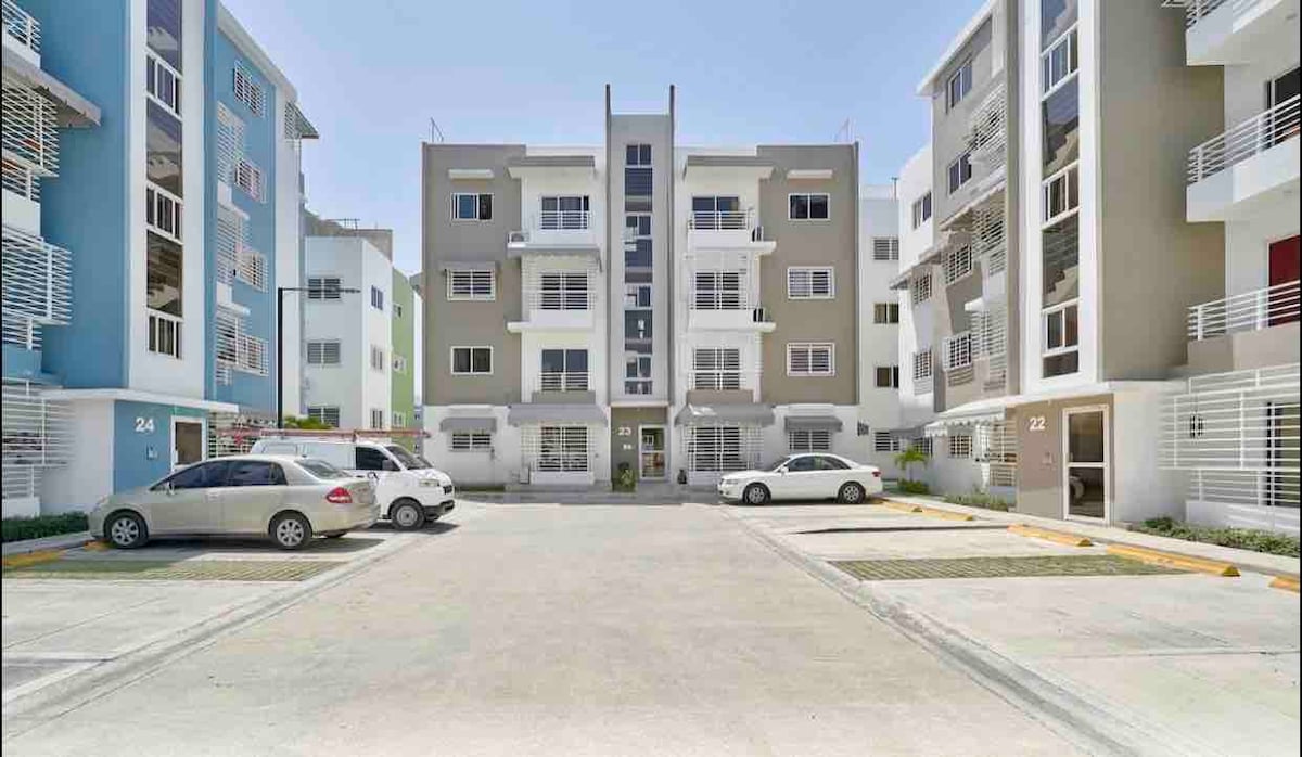 The image depicts a modern residential complex with multiple two-story buildings, featuring a central courtyard. Several parked cars are visible in front, with well-maintained pathways and green landscaping surrounding the entrance. The blue and beige façades of the buildings suggest a contemporary design.