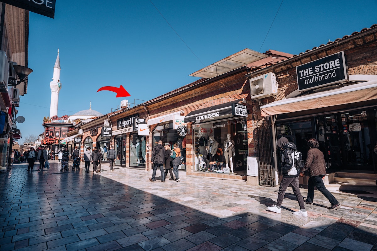 A bustling street scene in the main bazaar of Ohrid is depicted, featuring a mix of shops and pedestrians. The shops display a variety of brands, with people walking along a wide, cobblestone walkway under a clear blue sky. A prominent minaret stands in the background.