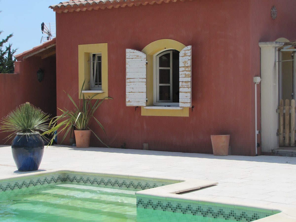 The exterior of the pool house is presented with a warm, terracotta facade and contrasting yellow accents. A window with decorative shutters is visible, alongside potted plants, including a tall, spiky plant. The pool area features pale tiles and inviting green water.
