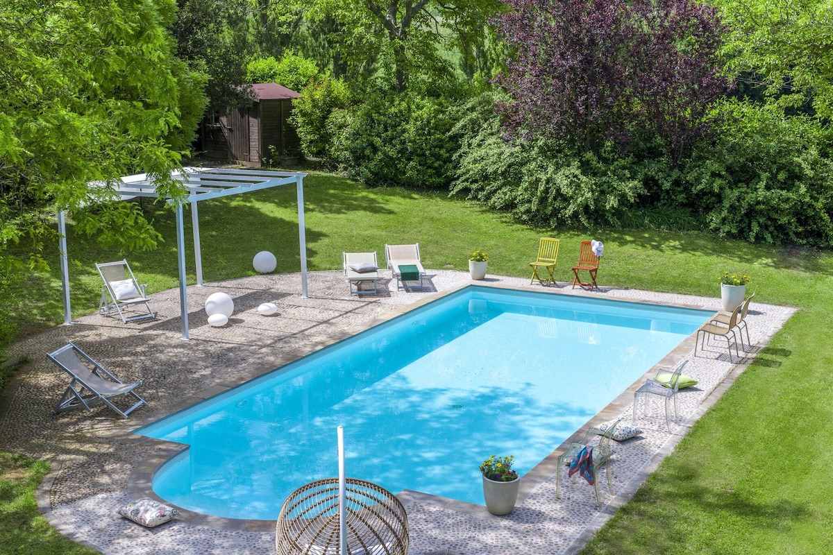 A private saltwater pool is set within a spacious garden, surrounded by greenery. Sun loungers and seating options, including a colorful chair and a hammock, are arranged around the poolside. A shaded area with a pergola provides a relaxing space for enjoyment.