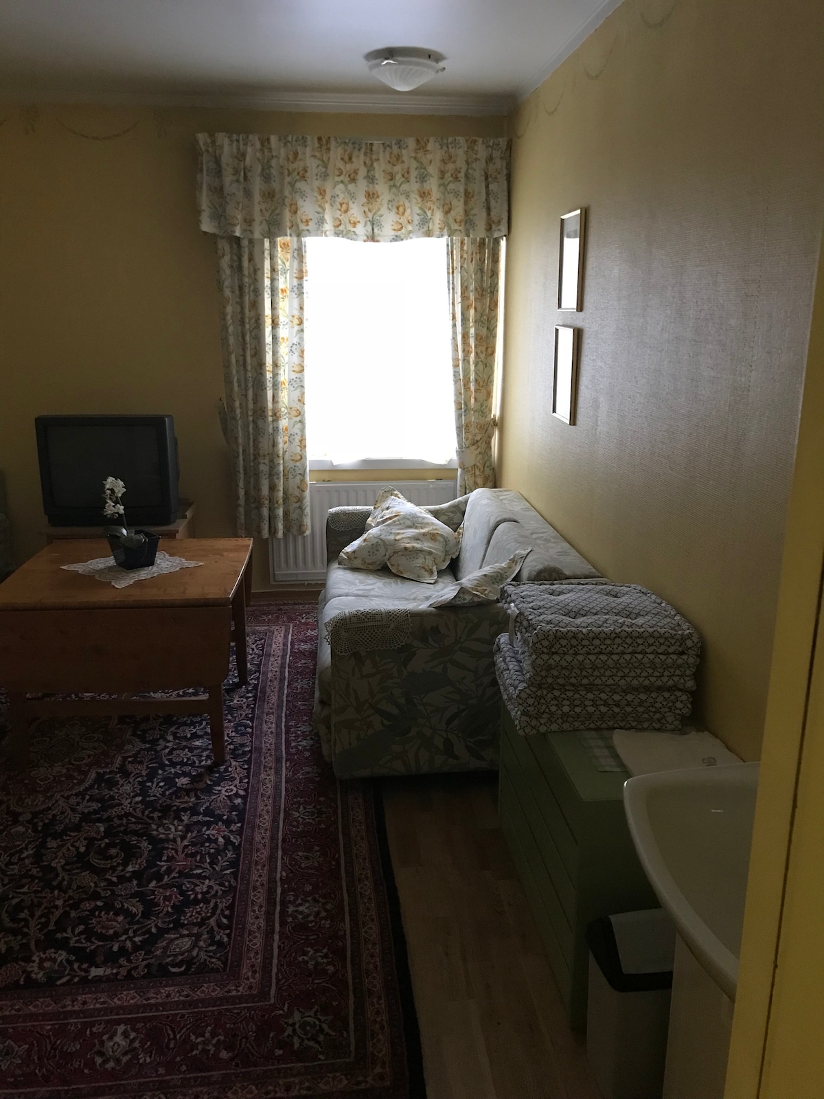 A cozy room is presented with a light yellow wall, featuring a window adorned with floral curtains. A comfortable sofa sits next to a small wooden table, and a television is positioned adjacent to a dresser. A patterned rug covers the wooden floor.