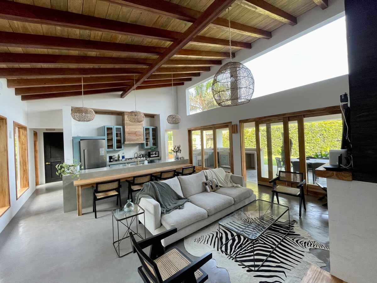 An open-concept living area is featured, showcasing high wooden beams and a spacious layout. Natural light pours in through large windows, illuminating the neutral tones of the furniture and décor. A cozy sofa is placed alongside a modern dining table, with a stylish rug enhancing the space.