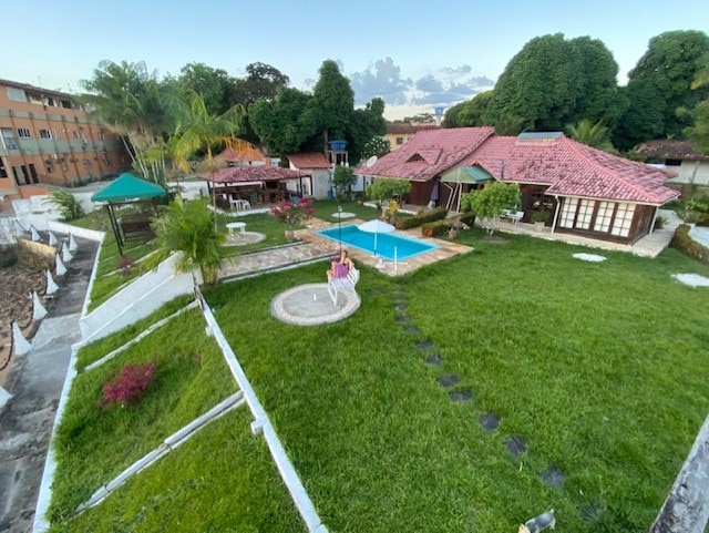 A spacious outdoor area features a lush green lawn, a swimming pool surrounded by decorative stones, and various seating options under shaded gazebos. Vibrant plants add color, while a charming house with a red-tiled roof completes the serene landscape.