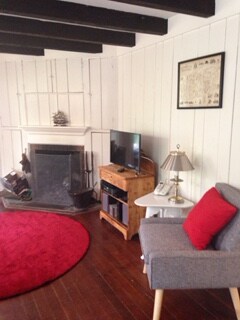 A cozy living area features a warm, circular red rug centered in front of a traditional fireplace. A wooden television stand holds a TV, while a small side table with a lamp complements a comfortable grey armchair with a red cushion.