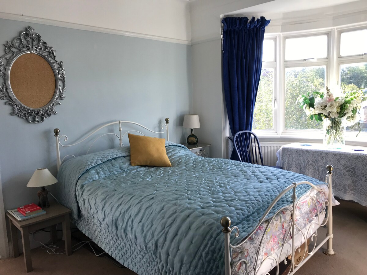 A spacious bedroom features a bed with a quilted blue coverlet and a decorative yellow pillow. A mirror with an ornate frame hangs above a nightstand, while a table dressed in a lace cloth is set by large windows with blue curtains, providing natural light.