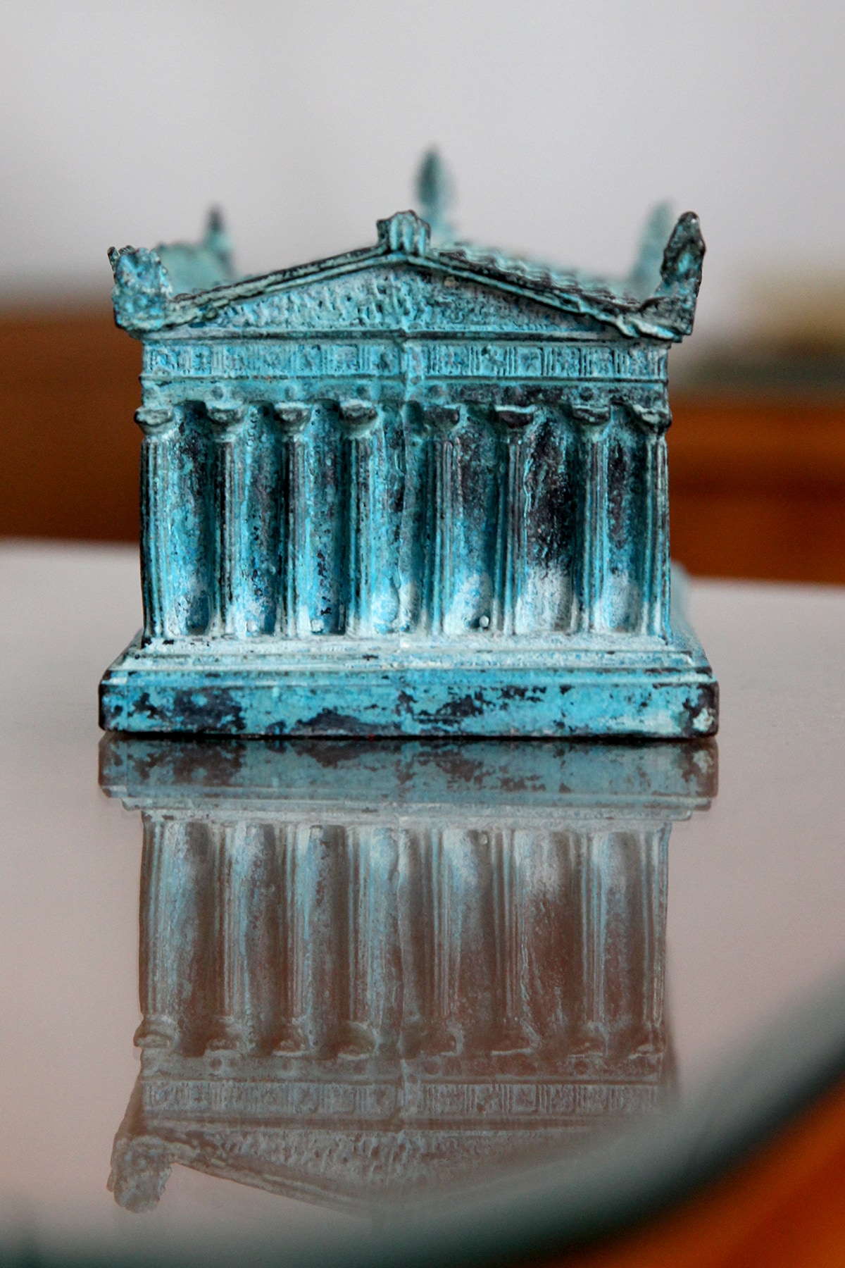 A detailed bronze replica of a classical building rests on a reflective glass surface. Columns and ornate features are visible, showcasing craftsmanship. The reflection enhances the object's presence, drawing attention to its intricate design and the subtle patina of aged metal.