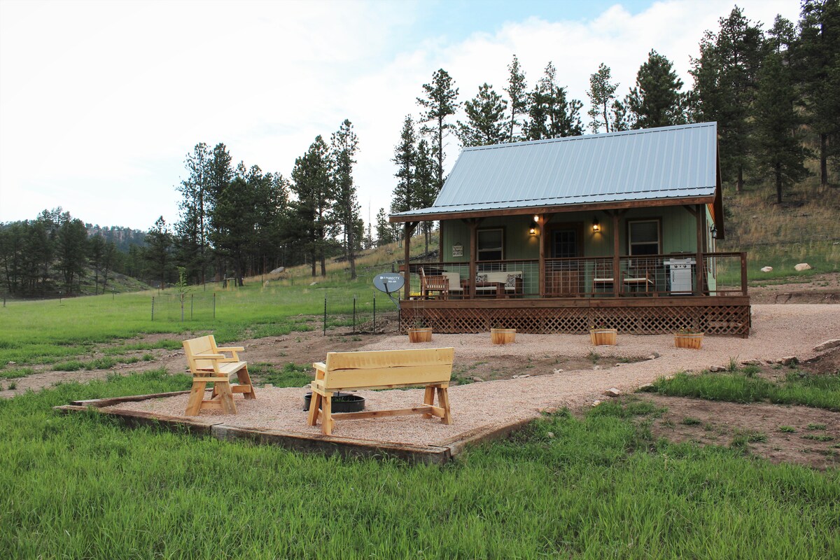 The farmhouse cabin is set against a backdrop of green hills, featuring a metal roof and wooden deck. Outdoor seating is arranged in front, with a fire pit nearby, offering a comfortable space to enjoy the surrounding nature.