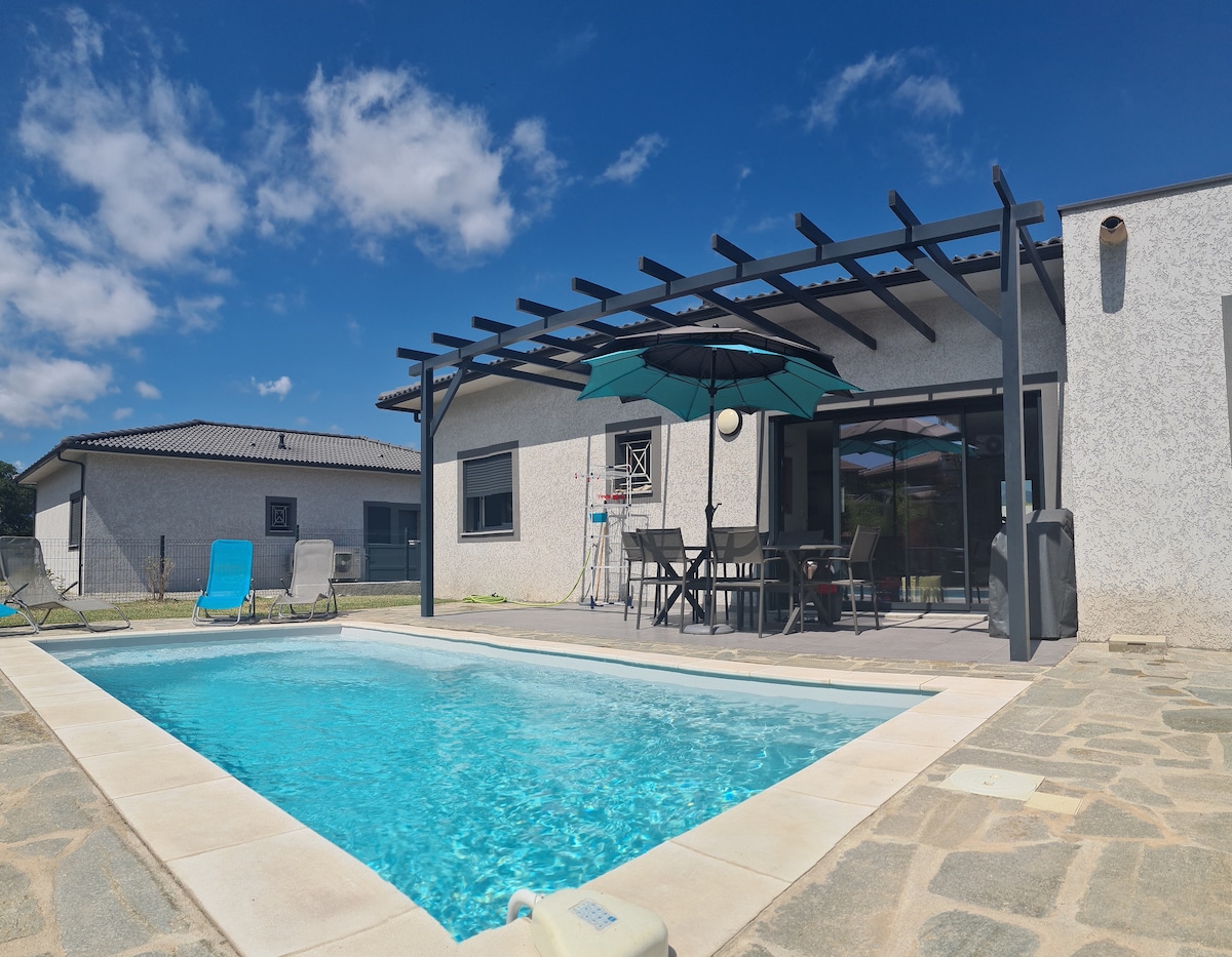 A modern villa is depicted, featuring a swimming pool surrounded by a paved area. A patio cover with an umbrella provides shade over a dining table and chairs. Sun loungers are placed nearby, and the clear blue sky adds to the serene atmosphere.