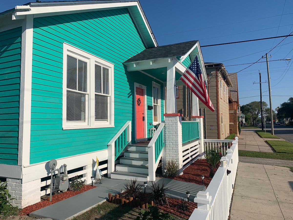 A charming bungalow with turquoise siding features a contrasting coral front door. An American flag is displayed outside, complemented by a well-kept garden with low-maintenance plants. The inviting porch is accessed by a set of steps, providing a glimpse of the neighborhood street.