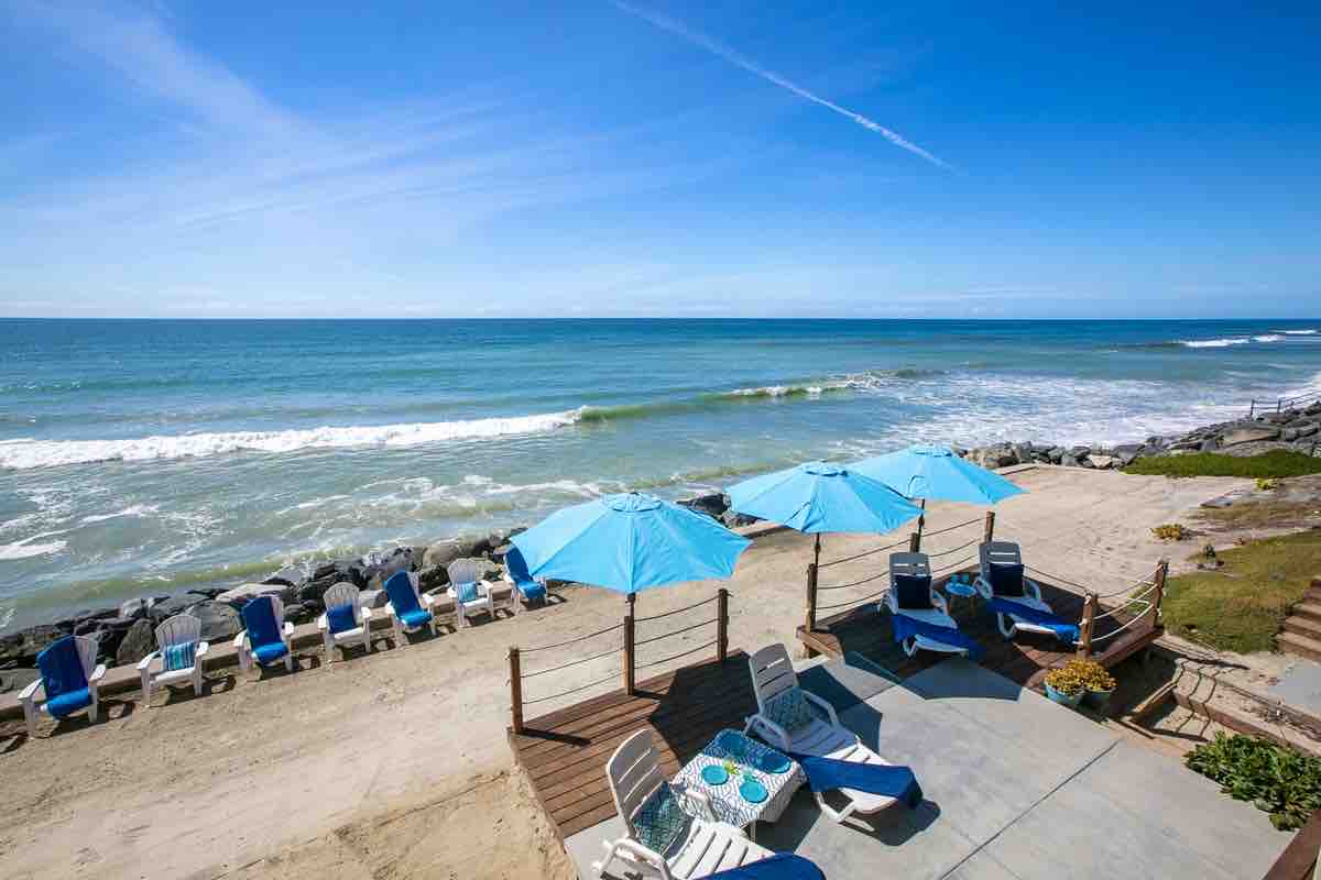 A spacious beachfront area is highlighted by several blue umbrellas providing shade over lounge chairs. Soft sand leads to gentle ocean waves, while a rocky barrier outlines the beach. The sky is clear with a few wispy clouds, enhancing the coastal landscape.
