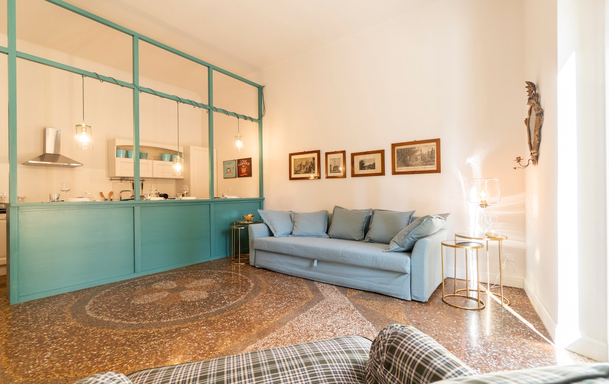 A spacious living area features a soft blue sofa, complemented by a decorative side table. Warm light fills the room, highlighting the patterned flooring and various framed pictures on the wall. The open kitchen area is visible in the background, showcasing modern design elements.