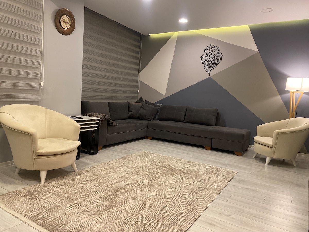 A modern living area features a spacious gray sectional sofa and two light-colored armchairs. A decorative wall art piece is centered above a subtle geometric design. A soft area rug covers the floor, and natural light is provided by a floor lamp.