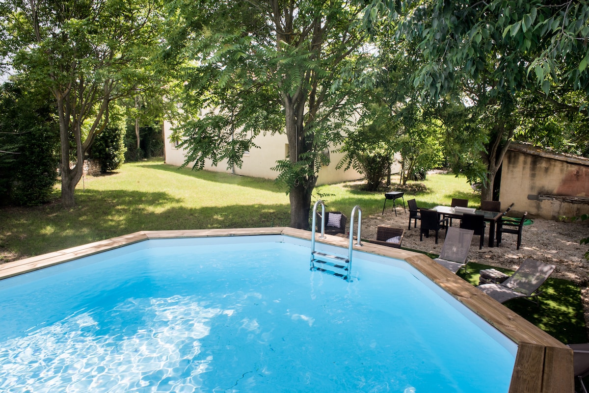A bright, inviting pool is surrounded by lush greenery and trees, providing shaded areas for relaxation. Nearby, a dining set and additional seating are visible on the patio, offering options for outdoor gatherings. The spacious garden is well-maintained, enhancing the tranquil outdoor setting.