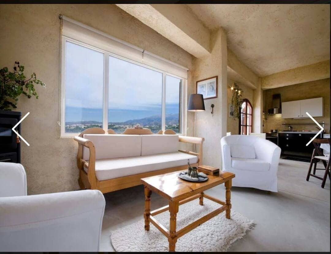 A bright sitting area is highlighted by a light-colored sofa and two white armchairs. A wooden coffee table is positioned in front, with a view of the sea visible through the large window. The space is open, showcasing a kitchen area in the background.