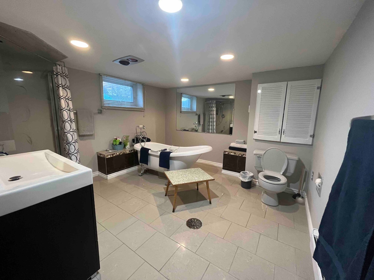 The spacious bathroom features a claw foot tub opposite a modern shower. A large mirror reflects soft lighting, while neutral-colored tiles provide a clean aesthetic. Storage cabinets are positioned alongside a dressing stool, adding functionality to the layout.